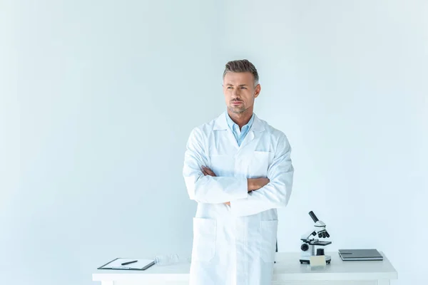 Cientista bonito de pé com braços cruzados perto da mesa isolado em branco — Fotografia de Stock