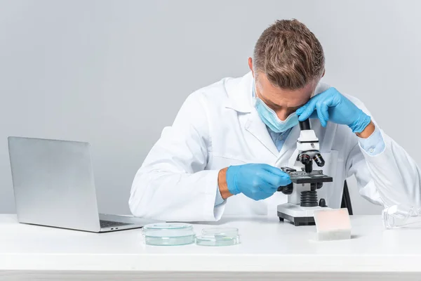 Guapo científico en máscara médica y guantes médicos mirando microscopio aislado en blanco - foto de stock