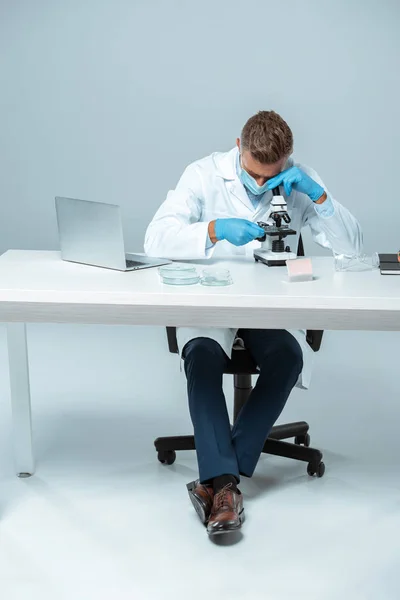 Químico bonito em máscara médica e luvas médicas olhando para microscópio isolado em branco — Fotografia de Stock