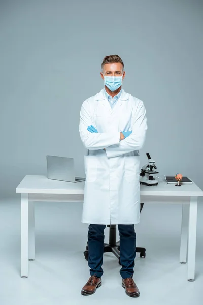 Guapo científico en máscara médica y guantes médicos de pie con los brazos cruzados aislados en blanco - foto de stock