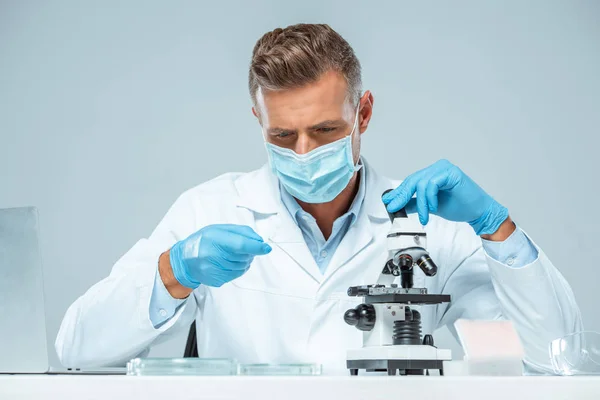 Guapo científico en máscara médica y guantes médicos haciendo prueba con microscopio aislado en blanco - foto de stock