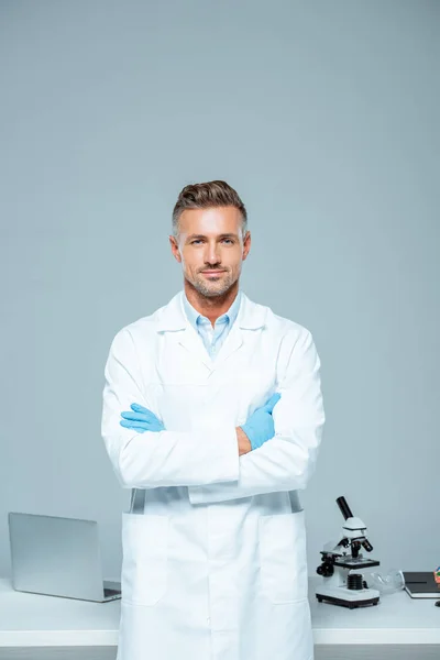 Handsome scientist in white coat and latex gloves standing with crossed arms and looking at camera isolated on white — Stock Photo