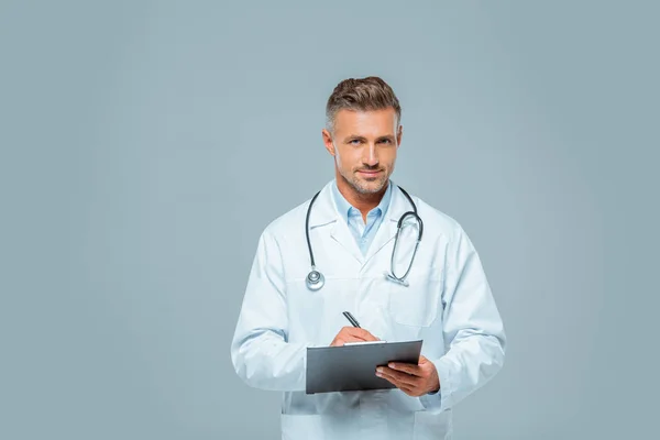 Handsome doctor with stethoscope writing something to clipboard and looking at camera isolated on white — Stock Photo