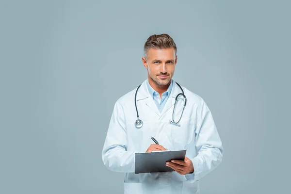 Guapo doctor con estetoscopio escribiendo algo al portapapeles y mirando la cámara aislada en blanco - foto de stock