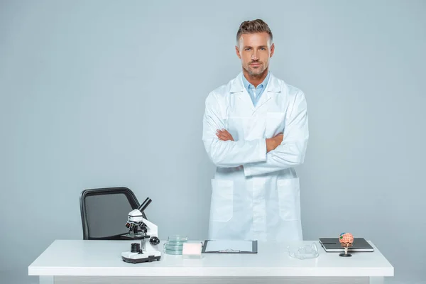 Bello scienziato in piedi con le braccia incrociate vicino al tavolo isolato su bianco — Foto stock