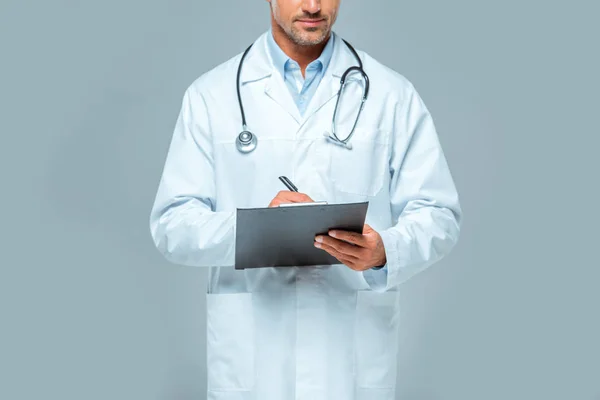 Imagen recortada de médico con estetoscopio escribiendo algo al portapapeles aislado en gris - foto de stock