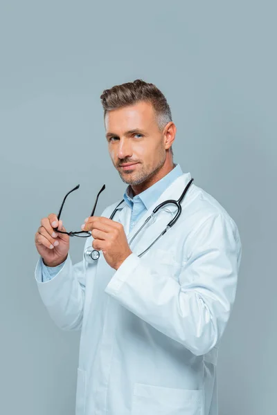 Bel dottore con stetoscopio che tiene gli occhiali e guarda la fotocamera isolata su grigio — Foto stock