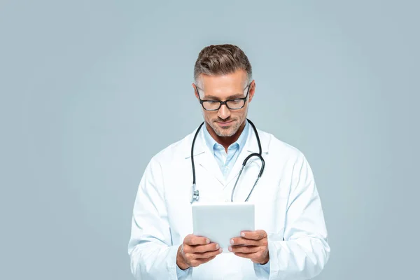 Guapo doctor con estetoscopio mirando tableta aislada en blanco - foto de stock