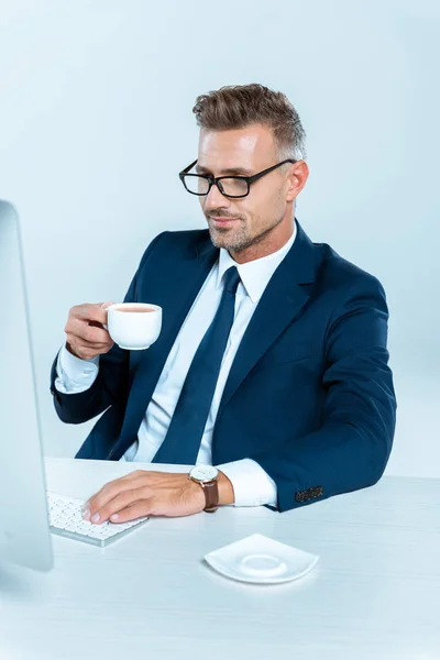 Bonito alegre empresário segurando xícara de café e olhando para o computador isolado no branco — Fotografia de Stock