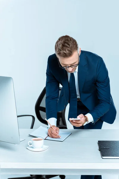 Gutaussehender Geschäftsmann schaut aufs Smartphone und schreibt etwas Isoliertes auf weiß — Stockfoto