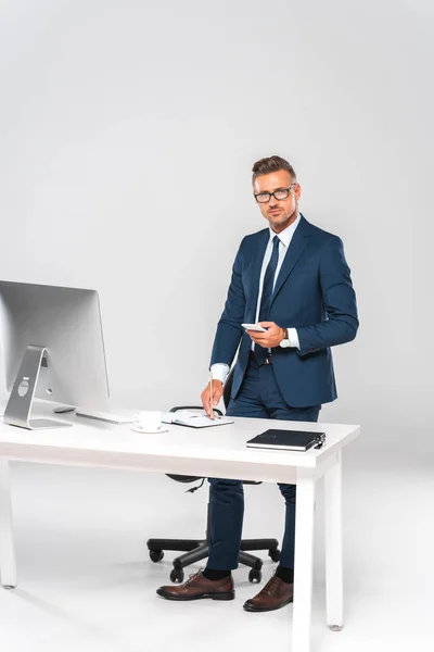 Guapo hombre de negocios sosteniendo teléfono inteligente y mirando a la cámara cerca de la mesa aislado en blanco - foto de stock