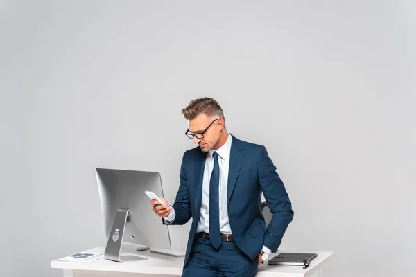 Homem de negócios bonito inclinado na mesa e usando smartphone isolado no branco — Fotografia de Stock