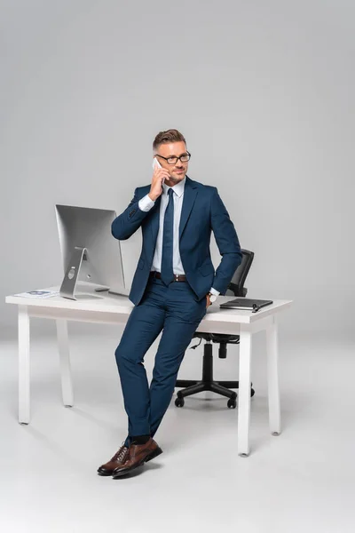 Guapo hombre de negocios apoyado en la mesa y hablando por teléfono inteligente aislado en blanco - foto de stock