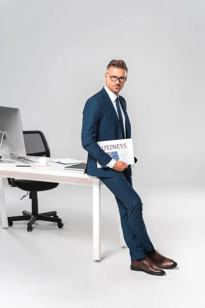 Guapo hombre de negocios sosteniendo el periódico, apoyado en la mesa y mirando a la cámara aislada en blanco - foto de stock