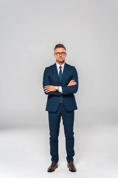 Hombre de negocios guapo en traje y gafas de pie con los brazos cruzados y mirando a la cámara aislada en blanco - foto de stock