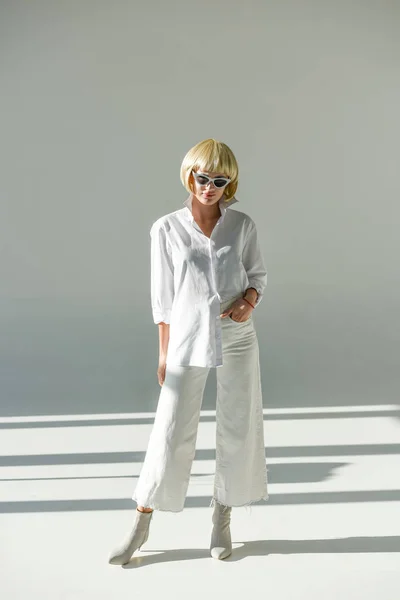 Attractive blonde woman in sunglasses and fashionable white outfit standing on white — Stock Photo
