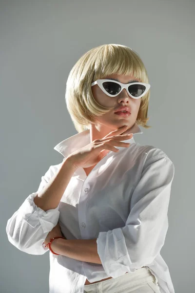 Jolie femme blonde en lunettes de soleil et tenue blanche à la mode touchant le menton et regardant loin isolé sur blanc — Photo de stock