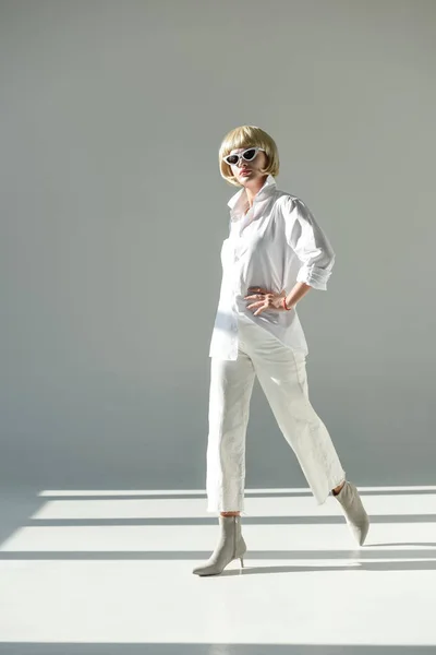 Jolie femme blonde en lunettes de soleil et tenue blanche à la mode marchant sur blanc — Photo de stock