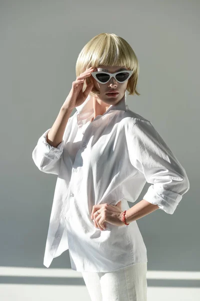 Beautiful blonde girl in sunglasses and fashionable white outfit standing with hand akimbo on white — Stock Photo