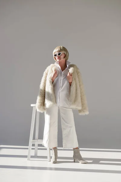 Attraktive blonde Frau mit Sonnenbrille und modischem Winteroutfit mit Kunstpelzmantel, die neben Stuhl auf weißem — Stockfoto