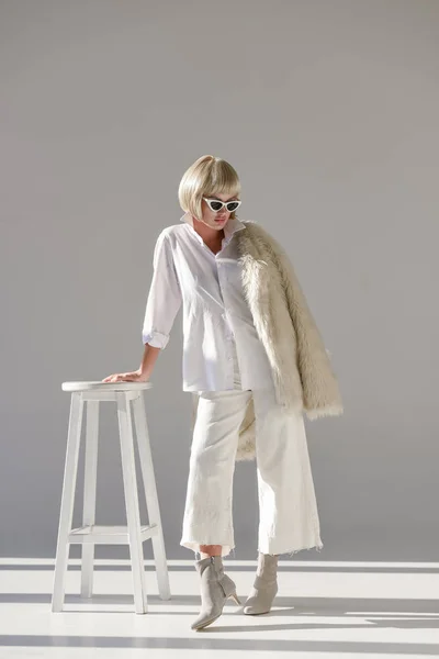 Attractive blonde woman in sunglasses and fashionable winter outfit with faux fur coat leaning on chair on white — Stock Photo