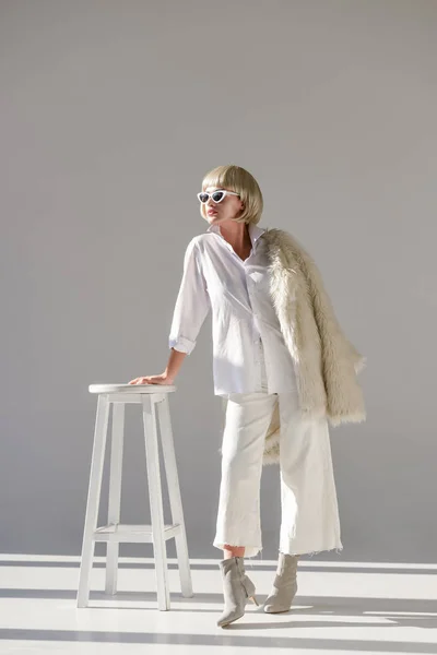 Красивая блондинка в солнечных очках и модный зимний наряд с искусственной шубой опираясь на стул на белом — стоковое фото