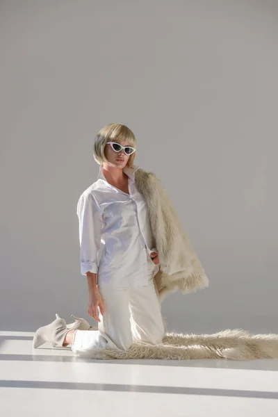 Привлекательная женщина в солнцезащитных очках и модном зимнем наряде с искусственной шубой, стоящей на коленях на ковре на белом — стоковое фото