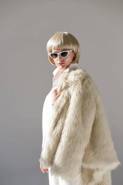 Portrait de jolie femme blonde en lunettes de soleil et tenue d'hiver à la mode avec manteau en fausse fourrure regardant la caméra sur blanc — Photo de stock