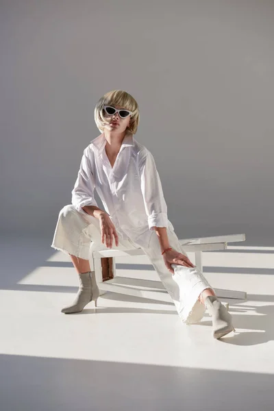 Jolie femme blonde en lunettes de soleil et tenue blanche à la mode assise sur une chaise sur blanc — Photo de stock