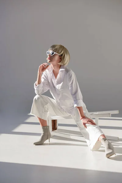 Attractive blonde woman in sunglasses and fashionable white outfit sitting on chair and looking away on white — Stock Photo