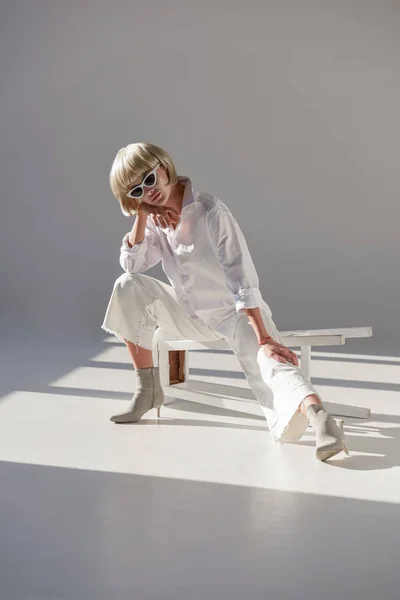Attraktive blonde Frau mit Sonnenbrille und elegantem weißen Outfit sitzt auf einem Stuhl auf weiß — Stockfoto