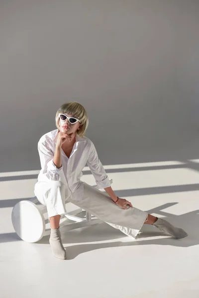 Atractiva mujer rubia en gafas de sol y traje blanco de moda sentado en la silla en blanco - foto de stock