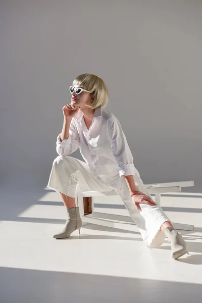 Jolie femme blonde en lunettes de soleil et élégant costume blanc assis sur la chaise et regardant loin sur blanc — Photo de stock