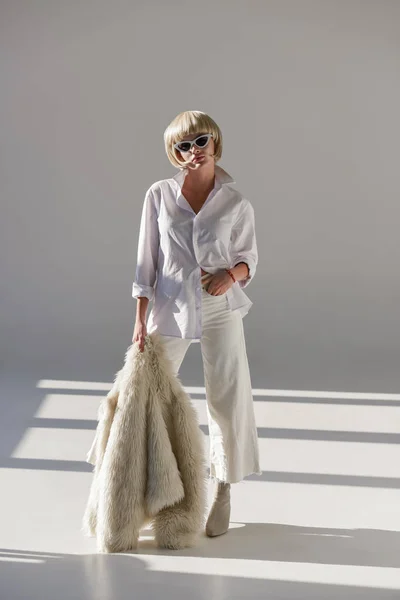 Attractive blonde woman in sunglasses and fashionable winter outfit posing with faux fur coat on white — Stock Photo