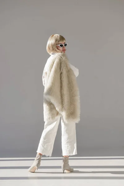 Back view of attractive blonde woman in sunglasses and fashionable winter outfit standing with faux fur coat on white — Stock Photo