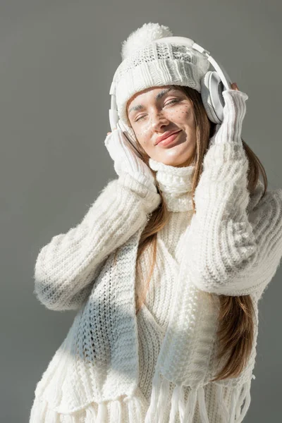 Happy attractive woman in fashionable winter sweater and scarf listening music with headphones isolated on grey — Stock Photo