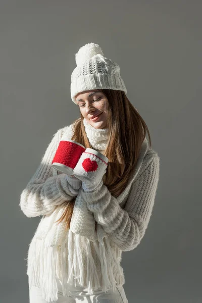 Весела приваблива жінка в модному зимовому светрі і шарфі тримає дві чашки ізольовані на сірому — стокове фото
