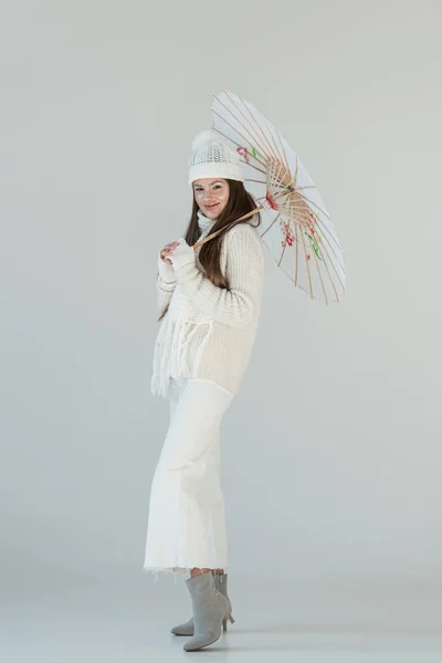 Привлекательная женщина в модном зимнем свитере и шарфе стоит с бумажным зонтиком и смотрит на камеру, изолированную на белом — стоковое фото