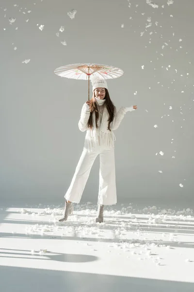 Щаслива жінка в модному зимовому светрі і шарфі, що стоїть під японською парасолькою, сніг падає на білий — Stock Photo