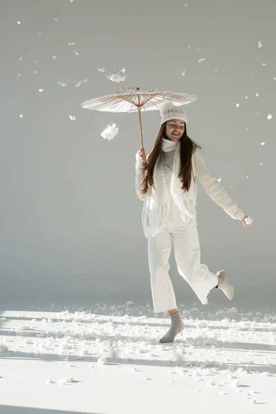 Attraktive Frau in modischem Winterpullover und Schal springt unter Papierschirm, Schnee fällt auf Weiß — Stockfoto