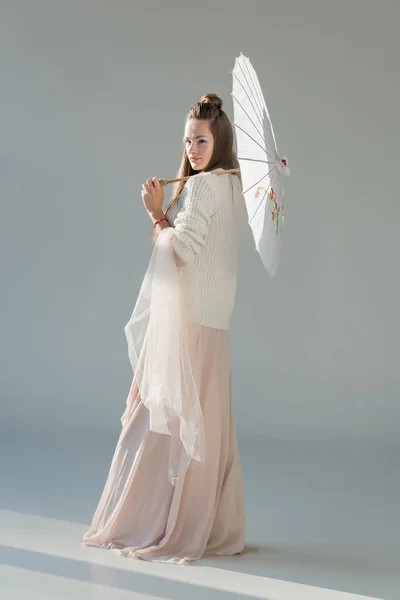 Vista lateral da mulher atraente na roupa de inverno elegante em pé com guarda-chuva de papel no branco — Fotografia de Stock