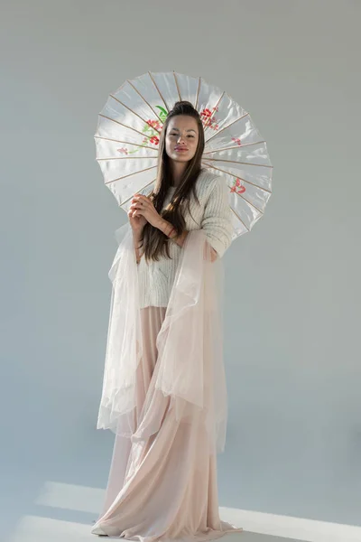 Attractive woman in fashionable winter outfit standing with japanese umbrella and looking at camera on white — Stock Photo