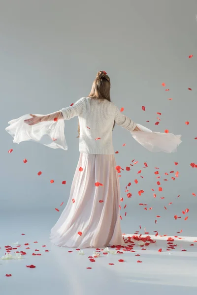 Rückseite der attraktiven Frau im modischen Winteroutfit, die sich unter fallenden roten Rosenblättern auf Weiß dreht — Stockfoto