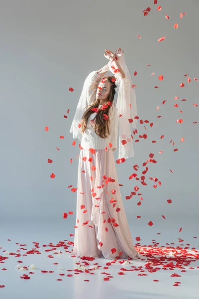 Attractive woman in fashionable winter outfit standing with closed eyes under falling red roses petals isolated on white — Stock Photo