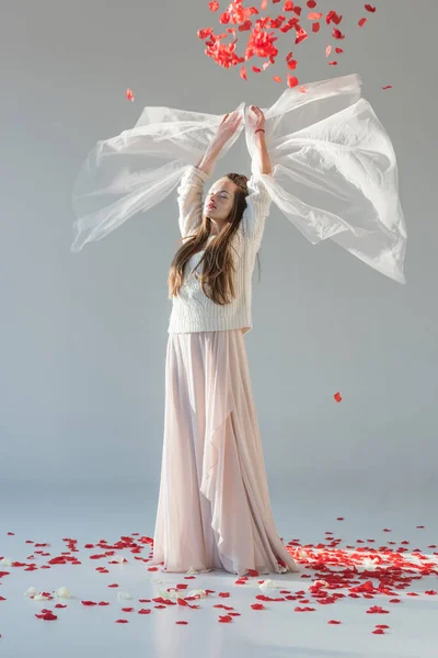 Jolie femme en tenue d'hiver à la mode debout avec les mains levées sous les pétales de roses rouges tombant isolé sur blanc — Photo de stock