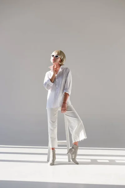 Belle fille blonde en lunettes de soleil et tenue blanche à la mode posant et regardant loin sur blanc — Photo de stock