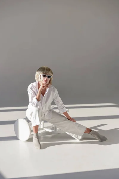 Attractive blonde woman in sunglasses and fashionable white outfit sitting on chair on white — Stock Photo