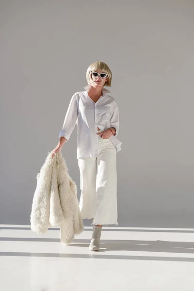 Jolie femme blonde en lunettes de soleil et tenue d'hiver à la mode marchant avec un manteau en fausse fourrure sur blanc — Photo de stock