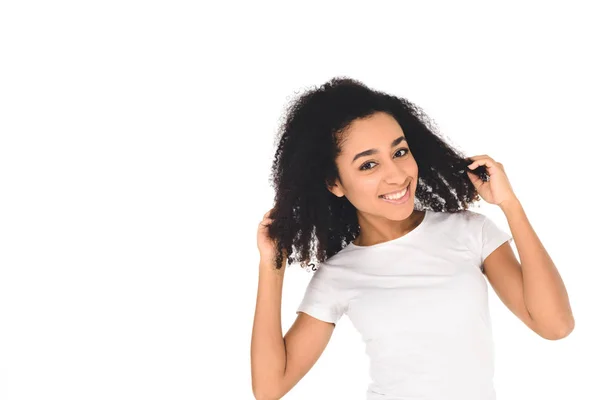 Atraente menina afro-americana tocando o cabelo e sorrindo para a câmera isolada no branco — Fotografia de Stock