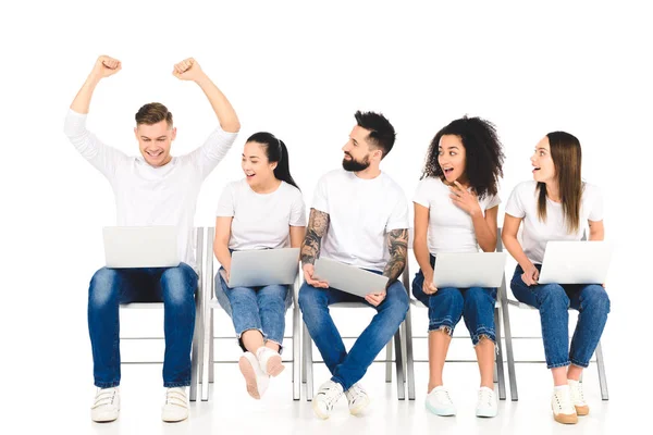 Handsome young man rejoicing with hands above head while multiethnic group of people using laptops isolated on white — Stock Photo